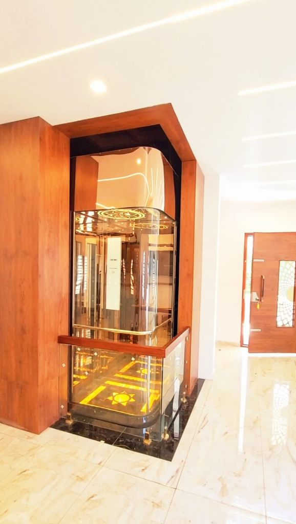 Curved glass home lift in a teak-clad surround, with a lit gold floor pattern and marble hall