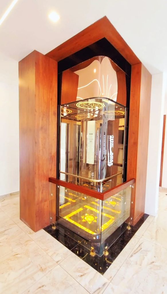 Curved glass lift with a gold ceiling and lit floor inside a teak surround